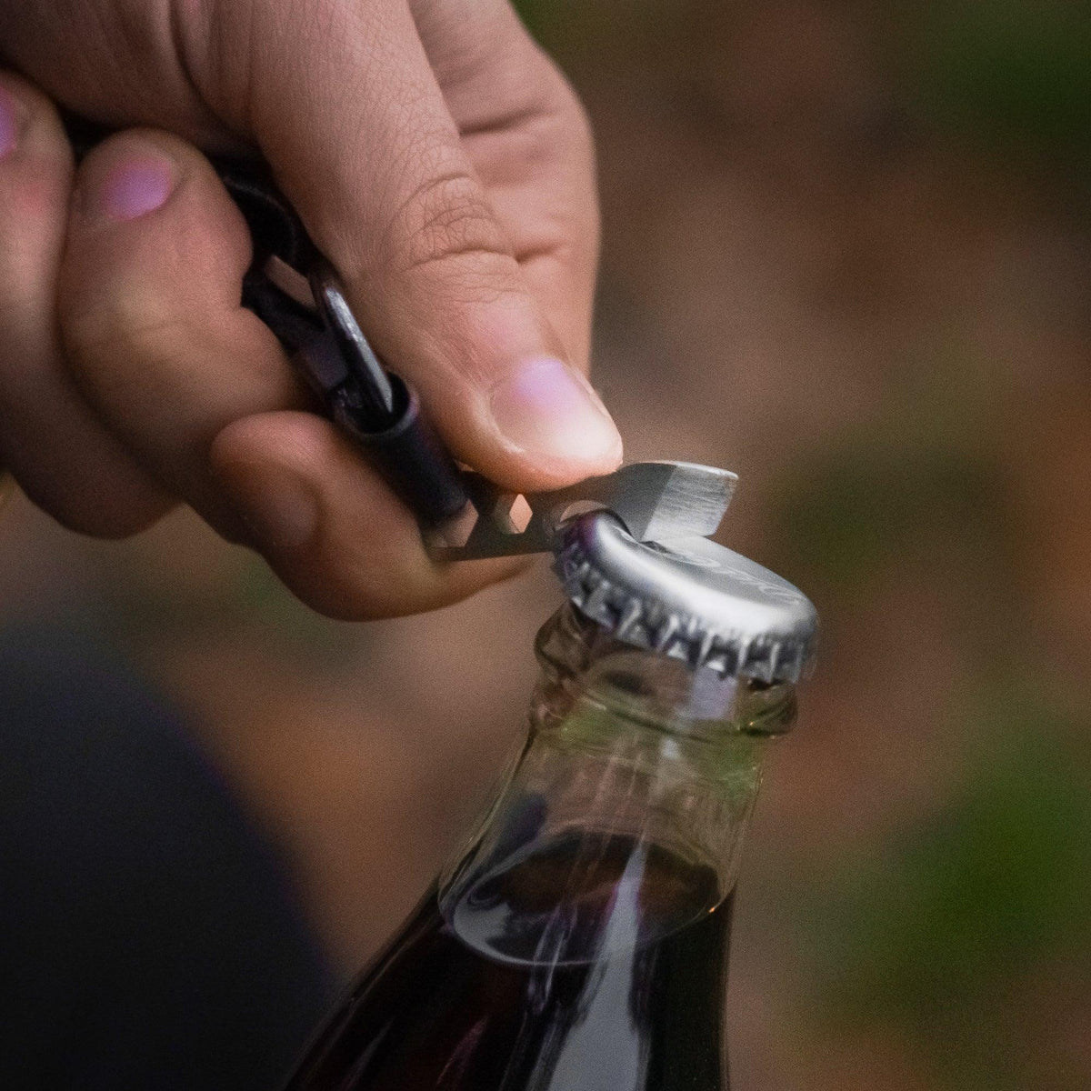Hand using compact multitool keychain bottle opener on soda bottle outdoors